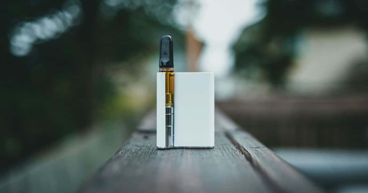 En vit e-cigarett med svart munstycke och synlig vätskebehållare står på ett träräcke utomhus.